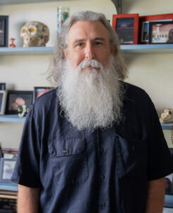 PHOTO: COM Professor Bernie Smiley poses for a portrait in his office, surrounded by personal belongings, photographs and more on shelves in the background.